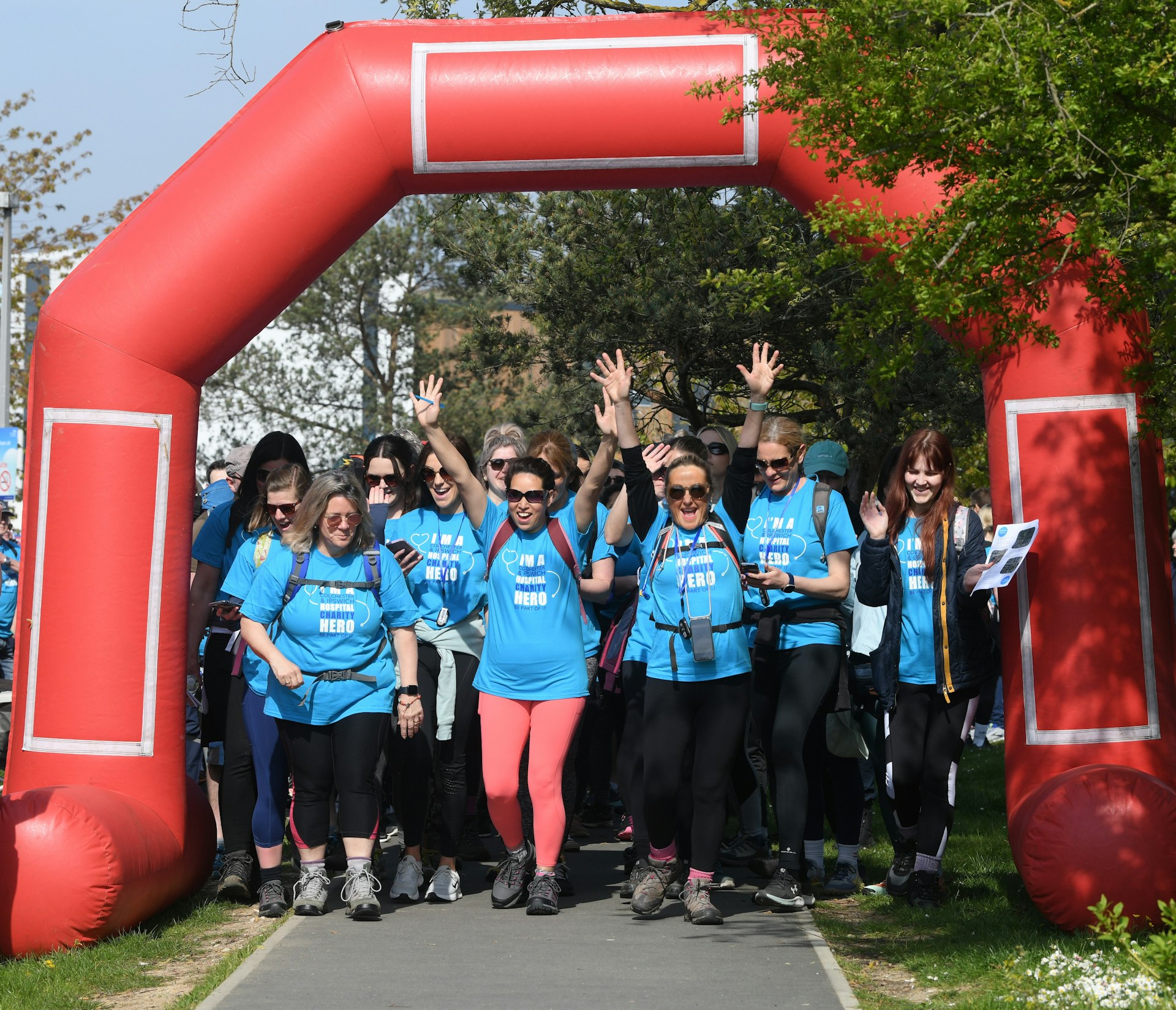 Group of walkers in blue Hospital Hero Hike t-shirts leaving the start line under a red archway